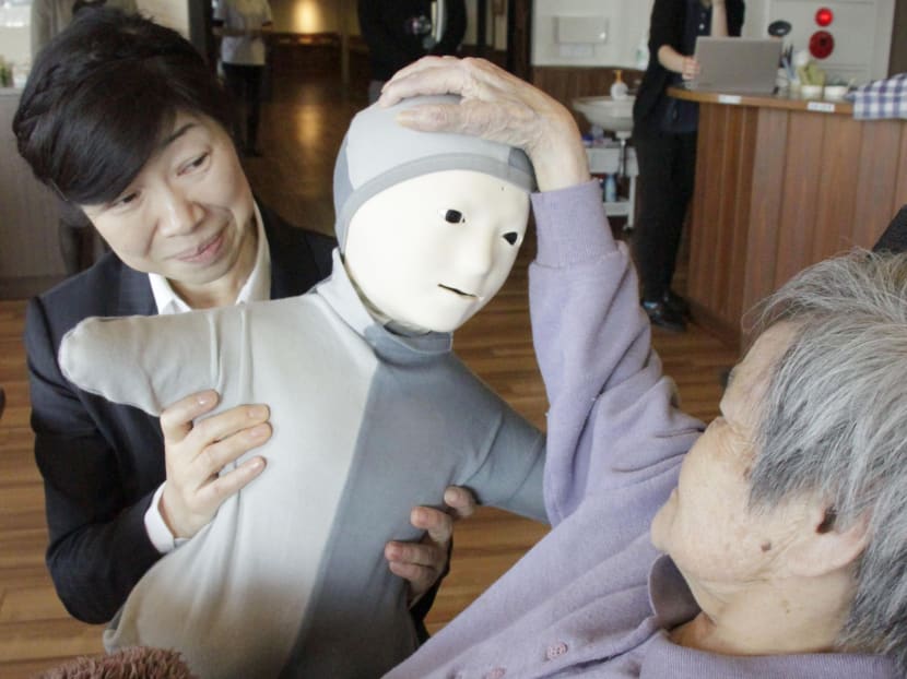 An elderly woman pats a Telenoid robot on the head at a nursing care facility in Natori, Miyagi Prefecture, northeastern Japan. A woman in the right back operates the robot. Photo: Kyodo News