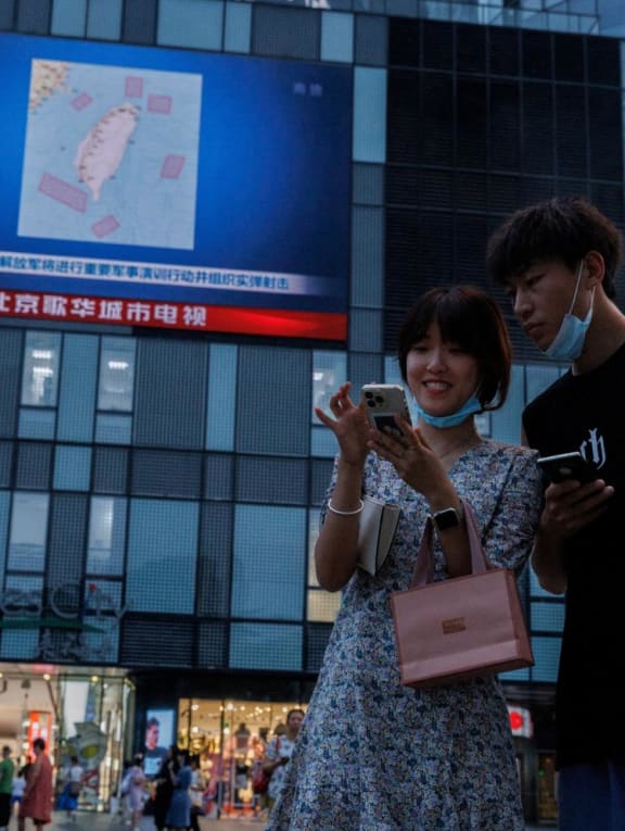 People stand in front of a screen showing a CCTV news broadcast, featuring a map of locations around Taiwan where Chinese People's Liberation Army (PLA) will conduct military exercises and training activities including live-fire drills, at a shopping center in Beijing on Aug 3, 2022. 