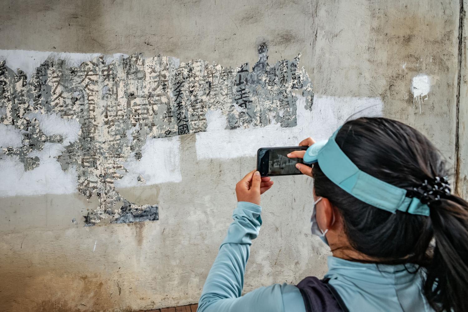 <p>A woman takes a photograph of some of the newly discovered work of the graffiti artist Tsang Tsou-choi on a wall under a railway bridge on Boundary Street in Hong Kong on May 28, 2022.</p>
