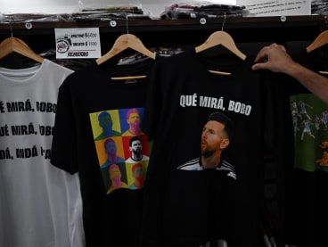 A man holds a t-shirt with an image of Argentine forward Lionel Messi and a phrase reading "What are you looking at, you fool?" at a store in Buenos Aires, on Dec 12, 2022.