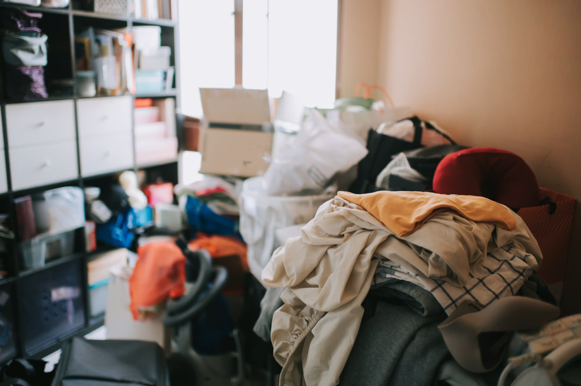 Clothes, bags, boxes and things piled high in a room, with shelving space also packed full of items.