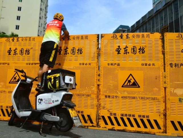 A courier stands on an electric bike to make a delivery over a barricade, amid lockdown measures to curb the coronavirus disease (Covid-19) outbreak in Sanya, Hainan province, China August 6, 2022. China Daily via REUTERS  