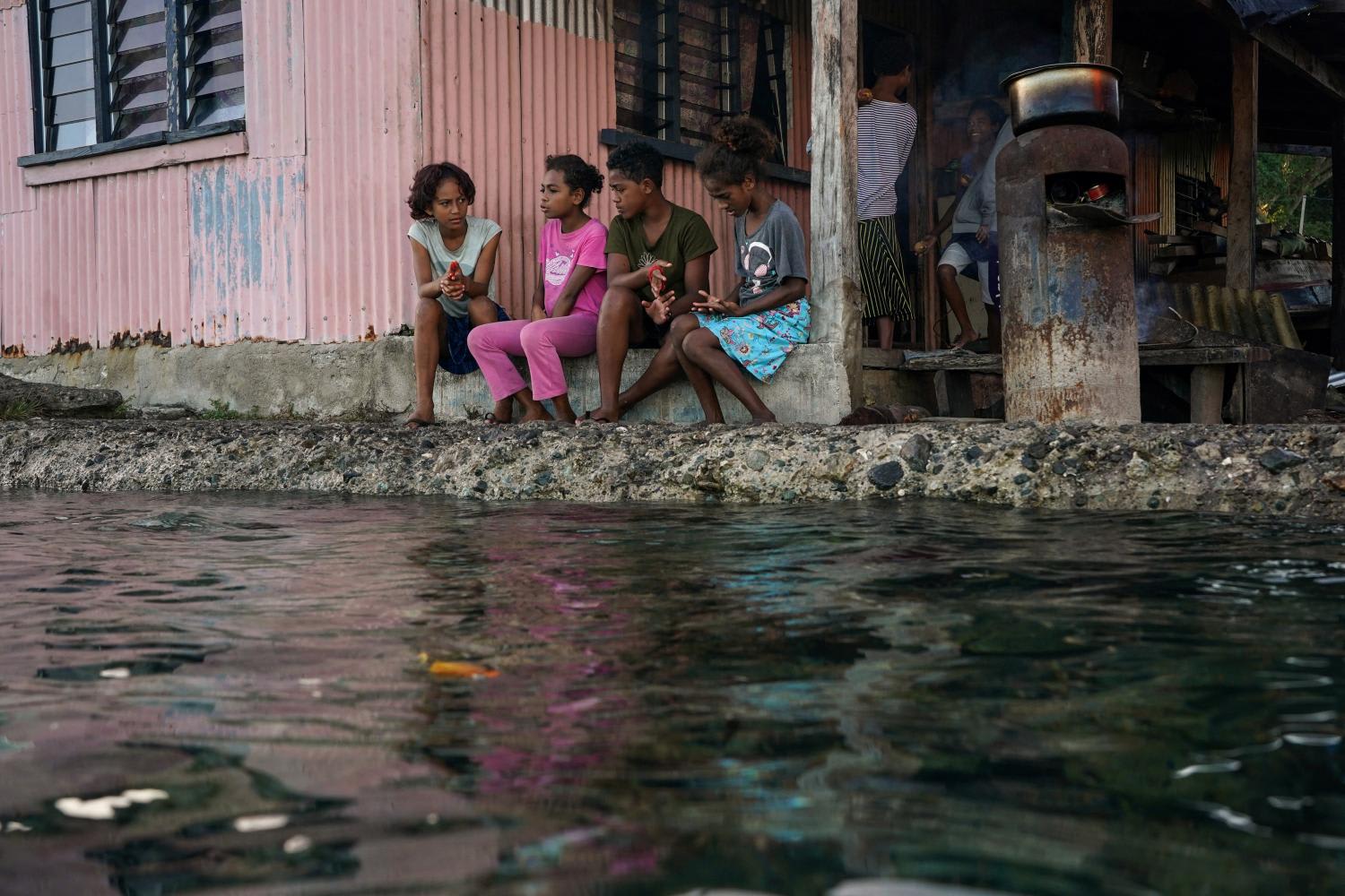 Children from a village in Fiji sitting outside their home next to a flooding sea wall at high tide. 